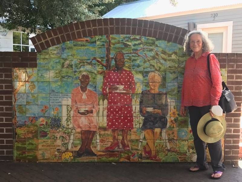 A picture of Jacqui Dorsey (the artist) standing next to a mural she painted featuring 3 the Eastside founding mothers: Artemisia Bowden, Mattie Briscoe, and Myra Hemmings