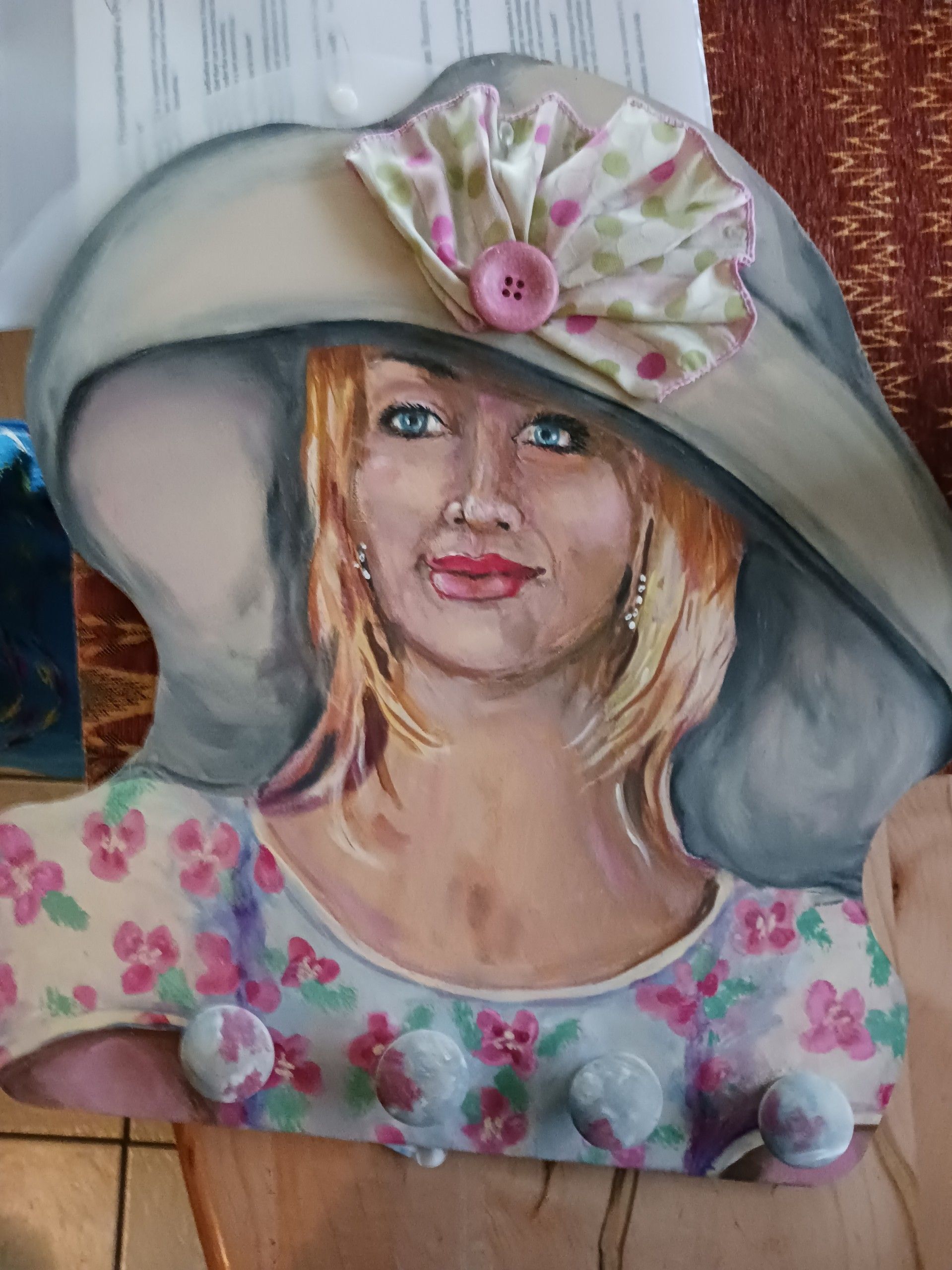 A blonde woman wearing a white hat and white and pink dress. The hat features a pink and green cloth accent, fastened via a pink button. Painted onto a wood known as jewelry head.